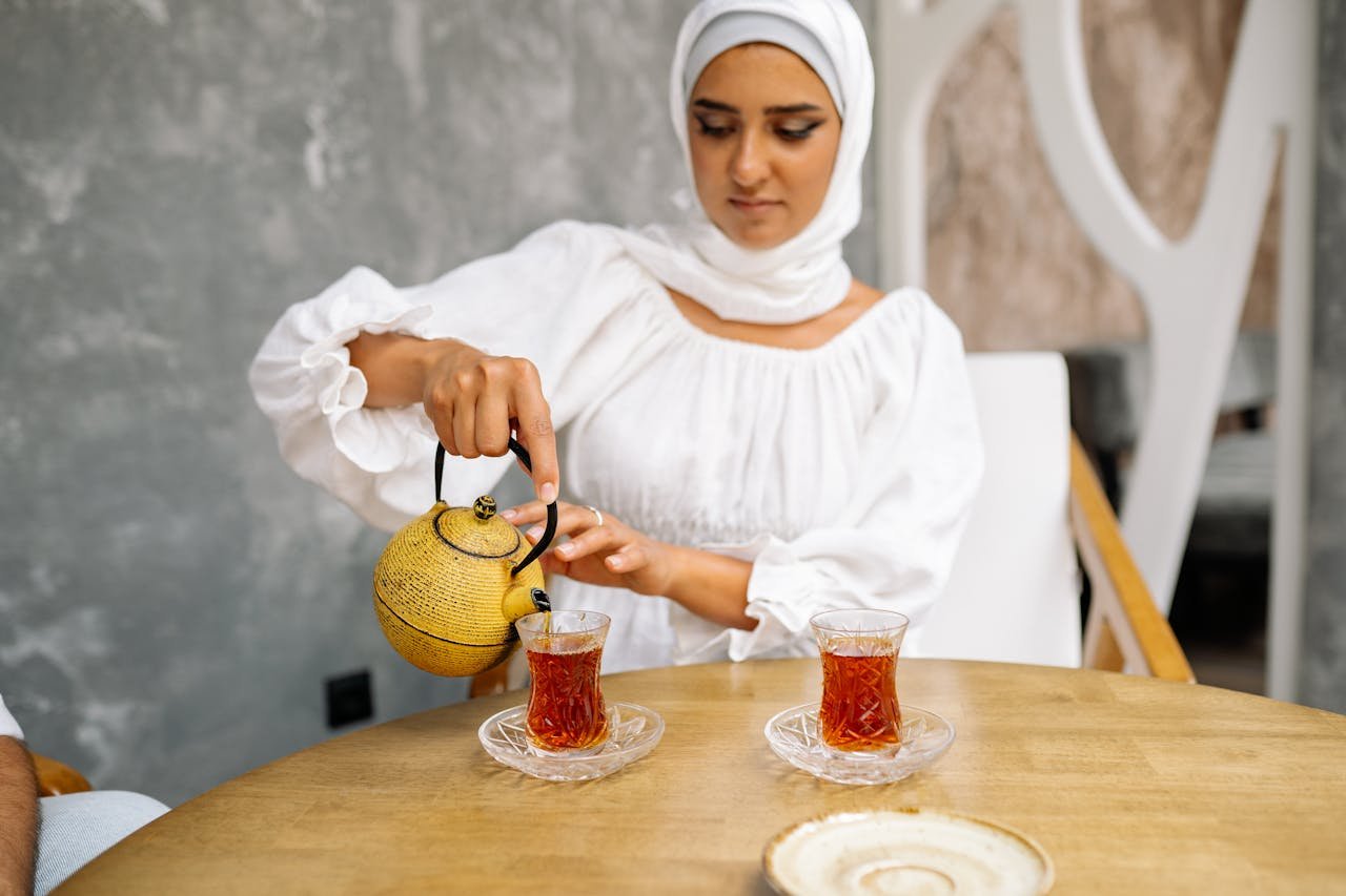 Woman in hijab pouring Turkish tea indoors, highlighting cultural traditions.